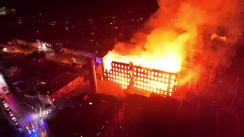 An aerial view of a large fire with flames and smoke coming out of a mill building in the middle of a town.