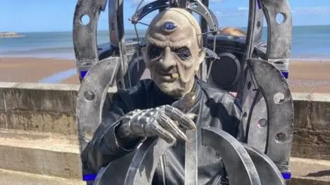 BBC/Carla Fowler A person dressed in a Doctor Who Davros character standing outside Scarborough Spa. The individual, who is wearing a semi-scary looking mask, a black leather jacket and a silver-robotic glove, is surrounded by metal-looking structures around their body.