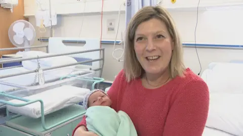 A baby wrapped in a green blanket is held by a blonde haired woman wearing  a red jumper