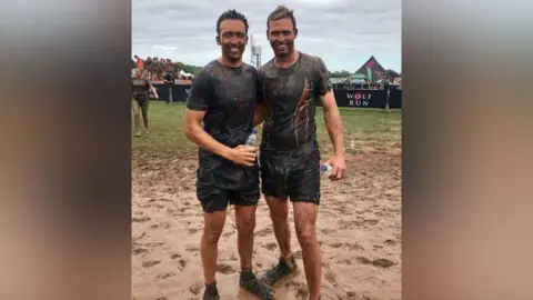 Two brothers standing arm in arm in a very muddy field that is saturated in mud. They are wearing black T-shirts with writing on that says "wolf run". they are covered in mud from head to toe and are smiling.