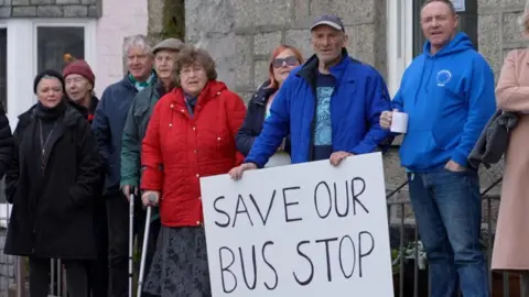 A group of Mousehole residents gathered in the village centre with one holding a placard saying 'Save Our Bus Stop' 