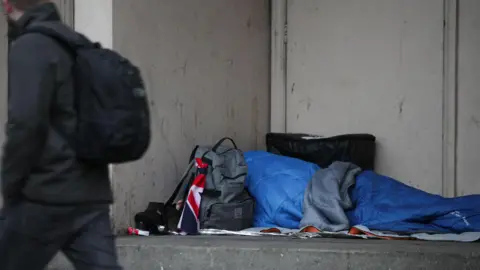 An unseen person lying in a doorway on the floor in a blue sleeping bag with a grey rucksack with a Union Jack flag tied around the handle. A person in a black coat and trousers with black rucksack glances as he walks past.