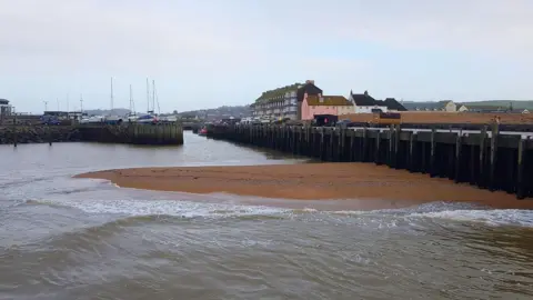 West Bay UK A large sand bar protrudes from the water in the outer harbour at West Bay. The harbour wall or east pier runs along the side of the photo. The photographer is standing on the west pier wall looking back towards the inner harbour which is full of small boats and lined with a terrace of Victorian-era buildings.