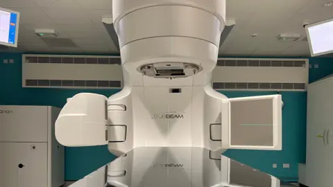 Dan O'Brien/BBC A large cream coloured radiotherapy machine sits in a room. It has a flat shiny table to lie in, with an overhead machine above it. On the ceiling are two computer screens. The logo says "TrueBeam".