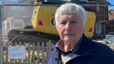Bryony Nierop-Reading, a woman standing in front of her house which is being demolished. She is looking directly at the camera.