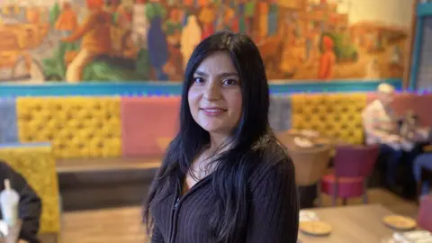 Image of Nisha Dhaka in an Indian restaurant, standing in front of a table. She has long dark hair and is wearing a ribbed dark brown zipper top.