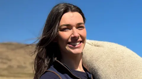 A young woman with long brown hair. She has a woolsack over one shoulder and is standing in front of grassland. The sky behind her is bright blue. 