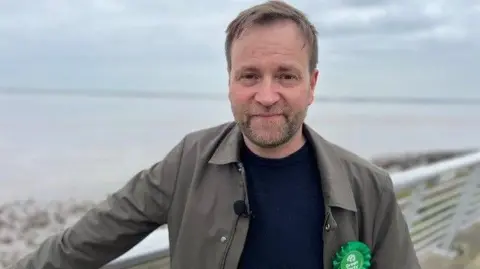 White male with short brown hair and stubble smiling into the camera. He is wearing a brown jacket, a blue jumper and he's standing in front of the Humber Estuary. He's wearing a Green Party rosette.