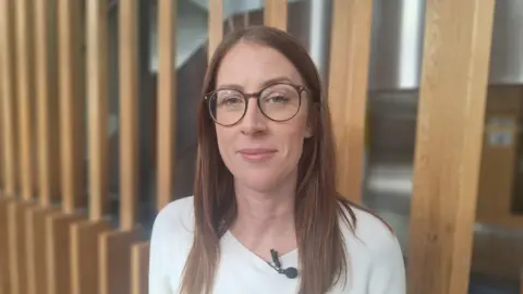 Sophie Legge is standing in front of a wall of wooden slats. She is wearing a white sweater and is looking into the camera with one eyebrow slightly raised. She has long brown hair and is wearing brown glasses.