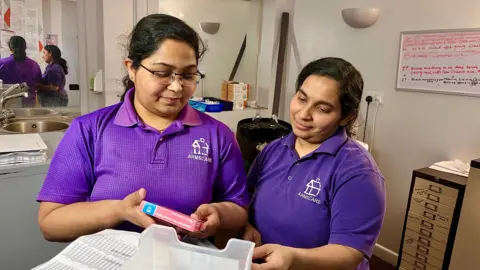 Two women wearing purple uniform polo shirts are standing in a care home office looking at patients' medicines.