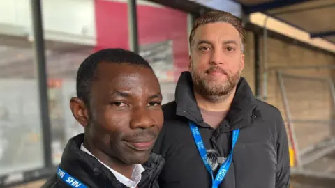 BBC Akin Afolabi and Mohammed Khan are pictured outside the Queen's Medical Hospital in Nottingham. They are wearing blue lanyards and black coats.