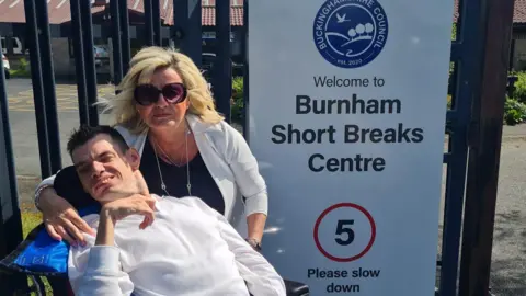 Hazel Howe Thomas Howe seated in a wheelchair beside a sign reading “Welcome to Burnham Short Breaks Centre,” with his mother standing behind with an arm around them; buildings and a car park are visible in the background under a partly cloudy sky.
