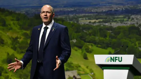 NFU Tom Bradshaw is standing on stage in a deep blue suit, with the NFU lectern behind him. His hands are open in an expressive gesture, as he addresses the conference. The stage back