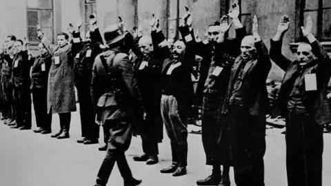 Corbis via Getty Images Inmates of the Warsaw Ghetto stand in line with their arms up as a German soldier walks towards them.