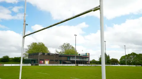 The rugby pitch at Norwich Rugby Club. The brick clubhouse is in the background. The white posts are in the foreground.