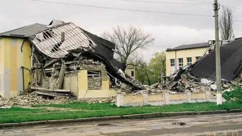 Volodymyr Rakov A large collapsed building next to a road. It appears to be a two-storey structure that has collapsed in the middle due to being struck.