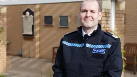 Avon and Somerset Police  Assistant Chief Constable Jon Cummins wearing a black police uniform with a blue police badge. He is standing in the courtyard of a light washed brick building and smiling at the camera. 