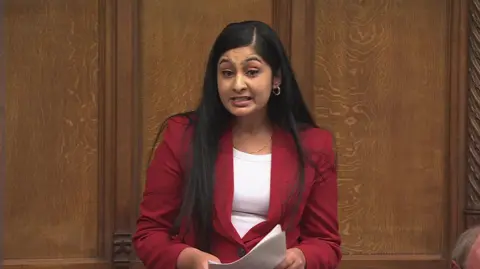 MP Zarah Sultana stood in the House of Commons wearing a red trouser suit and white top. She is holding A4 papers