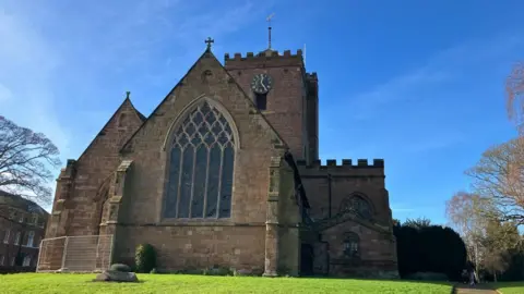 BBC Bright blue sky frames the background, with a stone church at the front of the picture and grass along the bottom. A few trees to either side of the church. 