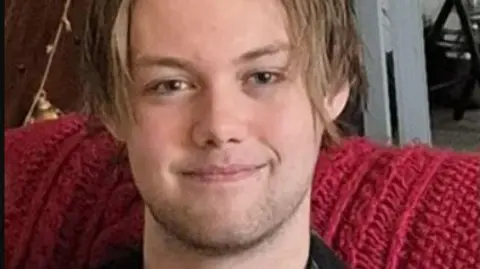 Ashton Kitchen-White smiles at the camera. He has light brown hair in a centre parting.