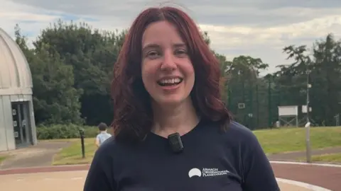 A woman with dark hair smiles at the camera, she is wearing a navy top
