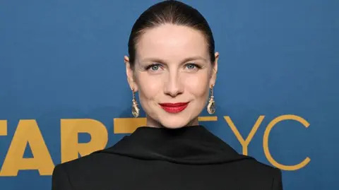 Caitriona Balfe, with her hair slicked back and wearing visible makeup including striking red lipstick as well as long jewelled earrings. She is standing in front of a blue sign with yellow writing on it.