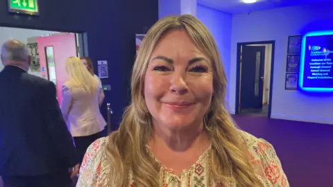 Lisa Evans has blonde hair smiling at camera. She is wearing a patterned white top with designs of red and black flowers. In the background is a blue neon sign.