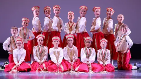 David Felton Photography A large group of girl dancers, dressed in cream and red national costumes, a number of them are kneeling down, the rest are standing. They are all smiling and looking straight at the camera. 