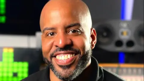 A man with shaved head and a trimmed black beard and moustache smiles at the camera. He is in a studio environment with audio and monitoring equipment visible behind him.