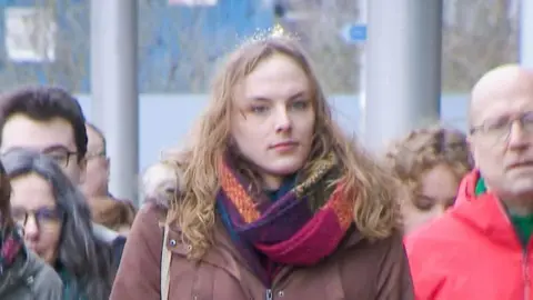 Dr Beth Upton - a trans woman with long blond hair, a multi coloured scarf and a beige coat, arrives with various supporters at a tribunal