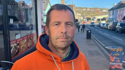 Terry Williams wearing an orange hoodie and stood next to a cafe.