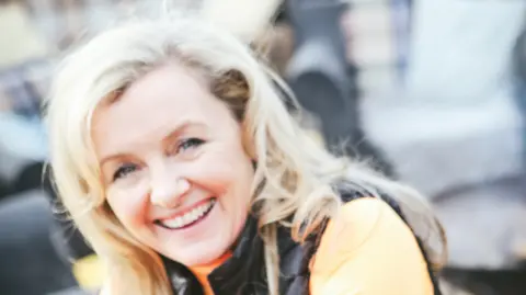 BBC Ruth Astbury looking at the camera. She has blonde hair and an orange top paired with a gilet.