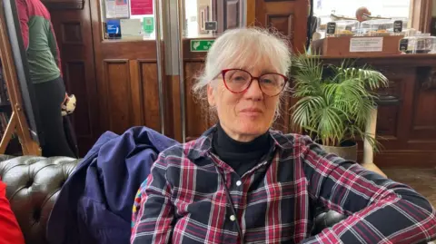 Vikki Irwin/BBC A woman is sitting on a leather sofa with her coat behind her. She is wearing a red and black checked shirt and a black polar neck underneath. She is wearing glasses and half smiling. 