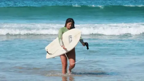 Mika Flower Surfer Mika Flower walking in sea holding a surfboard.