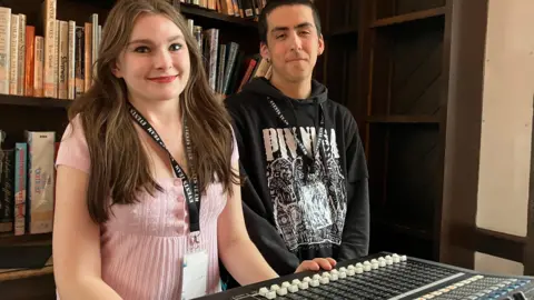 Jon Wright/BBC Cecilia Chard and Kieran Holland stand in front of bookshelves with a mixing desk in front of them.