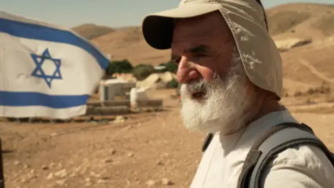 A man with a white beard and wearing a sun hat stands in front of an Israel flag