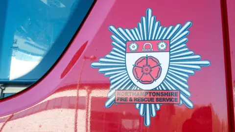 A close-up of the side of a red fire engine on which there is the emblem of the Northampton Fire and Rescue Service. 