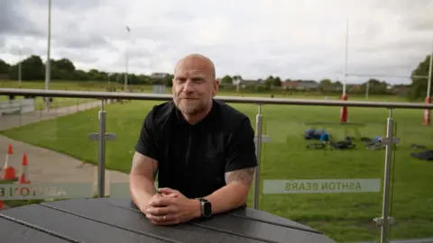 A man sits at a table with his hands clasped together, in front of a rugby pitch.