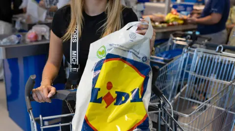 A woman pushes a shopping trolley away from a till in a supermarket. She is seen from the neck down. A number of other shoppers and trollies are in the background