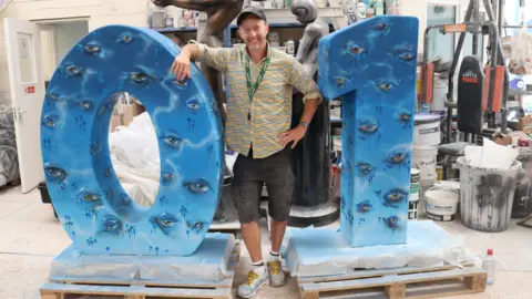 Alwynphoto Artist Spike stands with his scultpure of a number zero and number one painted blue and with eyes all over them.
