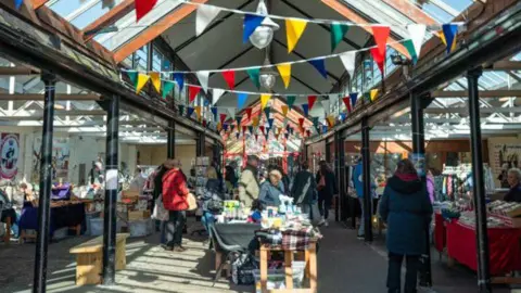 Torridge District Council A market scene is pictured, with flags handing over a long table of goods and people mingling around stalls as the sun casts a shadow from above.