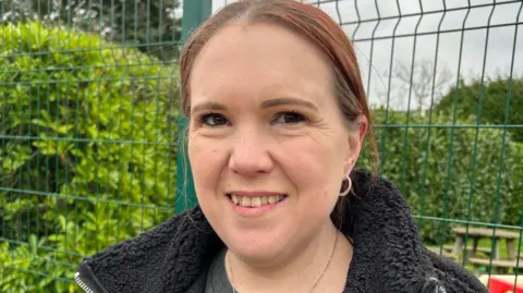 A woman with ginger hair and an ear ring, with a black fleece on, standing in front of a metal fence and green bushes