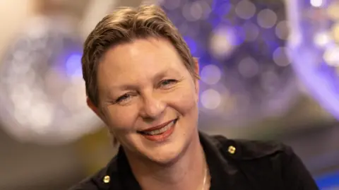 PA Media Dame Lucina Hackman is smiling widely at the camera. She has short, brown hair and is wearing a dark jacket. There are blurred lights behind her