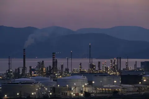 A view of the Motor Oil refinery in Agioi Theodoroi near Corinth, Greece during the evening. Behind the refinery, the sea and hills are visible but dark.