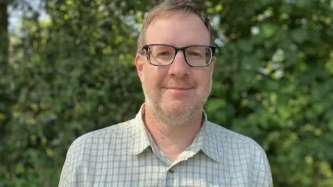 Man with short brown hair and moustache and beard and wearing glasses smiling at the camera. He is wearing a checked shirt that is Forestry Commission workgear. 