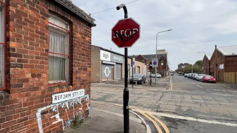 A street sign reading Reform Street is attached to a red brick wall containing graffiti. Next to the wall is a stop sign. 