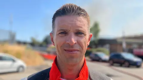 Shariqua Ahmed/BBC Rob Foreman has short grey hair and is wearing an orange hi-vis jacket. The background is blurred but there are several buildings and cars parked on the road.