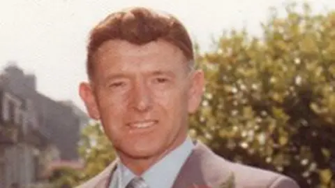 George Murdoch looking directly at the camera. He has dark eyes and brown hair and is wearing a grey suit with a light blue shirt.