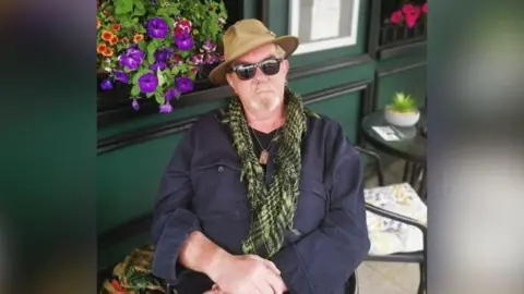 FAMILY HANDOUT A man sat outside a bar or restaurant. He is wearing a navy shirt, a green chequered scarf, dark sunglasses and a brimmed hat. He is beside some colourful flowers in a window box.   