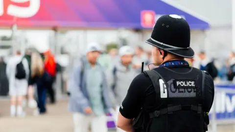 A police officer in a black T-shirt and black police helmet stands with his back turned and arms crossed among attendees at an event at Silverstone Circuit